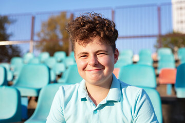 Teenager with brown, curly hair sits on the stand of a school stadium, looks at the camera and smiles. Sunset portrait.