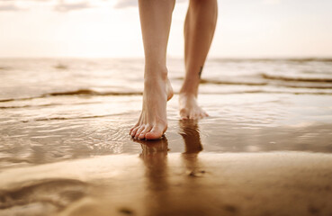 Close-up young woman's legs walking on a wave of sea water and sand on the beach. The concept of...