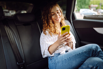 Modern woman in casual clothes sits in the back seat of a car, looks away, uses a mobile phone. Business, technology and travel concept.