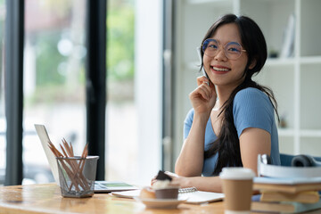 Asian woman sitting happily at work smiling relaxing and chatting with friends on social media search for information on laptop take notes in a notebook while sitting on a desk in his office at home