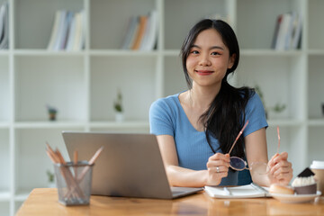 Asian woman sitting happily at work smiling relaxing and chatting with friends on social media search for information on laptop take notes in a notebook while sitting on a desk in his office at home