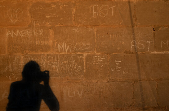 Background and texture of an old historical wall made of hewn stones. Tourists have scratched names or sayings on the wall. The shadow of a woman photographing the wall at sunset.