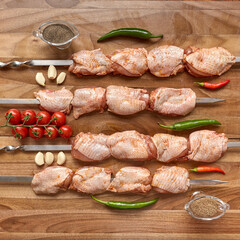 Metal skewers with raw uncooked chicken meat for frying on the wooden board with peppers near it, top view flatlay