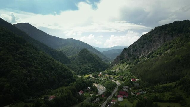Aerial drone shot of village houses in the valley surrounded by mountain range on all sides in Lepsa, Romania on a cloudy day. Green vegetation.