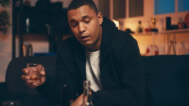 Drunk African American man holding glass with alcohol, sitting hungover on couch