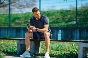 Naklejka premium Resting after fitness. Young sportsman sitting on bench and checking his phone after exercising outdoor.