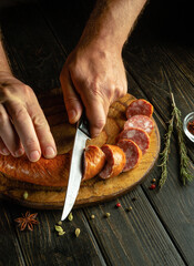 The cook cuts meat sausage with a knife on a wooden cutting board. The concept of preparing delicious sandwiches for breakfast on the kitchen table or fast food