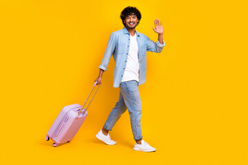 Full length photo of friendly man with wavy hair wear jeans shirt pull baggage say hi go empty space isolated on yellow color background