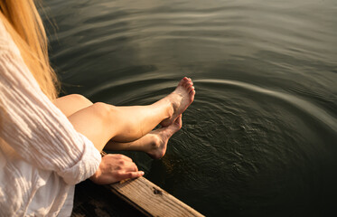 A woman relaxes by the lake, sitting on the edge of a wooden jetty, swinging her legs of the water.