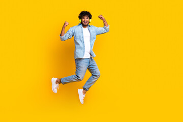 Fototapeta na wymiar Full length photo of cheerful lucky man wear jeans shirt jumping rising fists enjoying music empty space isolated yellow color background