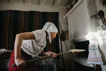 A woman in Ukrainian national dress works near a wood-burning stove. The traditional process of baking bread at home.