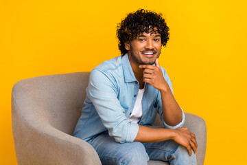 Portrait of good mood smart man with wavy hairstyle wear jeans shirt sit in armchair hand on chin isolated on yellow color background