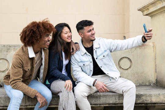 Three young diverse people taking a selfie with a smartphone sitting on a bank.Multiracial group smiling and taking a picture with a phone in a stone park bank.