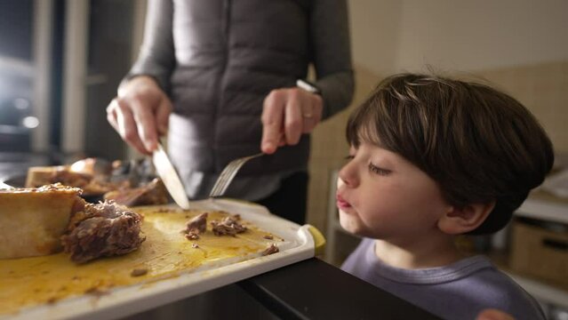 Little boy grabbing food by kitchen counter while mother prepare meal for family supper at night. Child observing mom cook food while grabs piece of meat