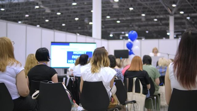 Speech of female employee at business conference for office workers. Speaker talks to audience at scene in crowded hall. Adult woman explains motivation to group of people. Summit meeting at modern