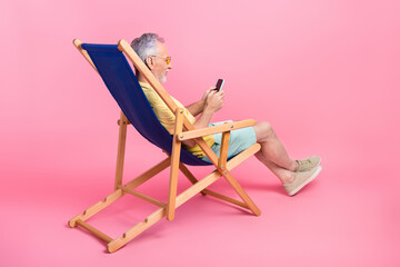 Full body photo of pensioner old man using his smartphone while lying sunbed vacation buy tickets plane isolated on pink color background