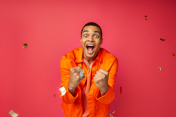 overjoyed african american man in orange shirt shouting and showing win gesture near confetti on red