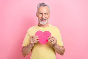 Photo of good mood kind man with gray beard dressed yellow t-shirt hands hold heart postcard on chest isolated on pink color background