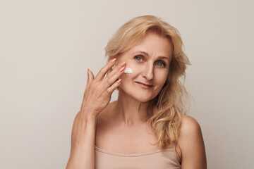 Portrait of luxurious looking 50s middle-aged mature blond woman smiling applying cream on face isolated on white studio background. Anti-aging skin care concept