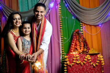 Indian family in ethnic wear celebrating Diwali with firecrackers at home with ganesha Statue in behind - obrazy, fototapety, plakaty