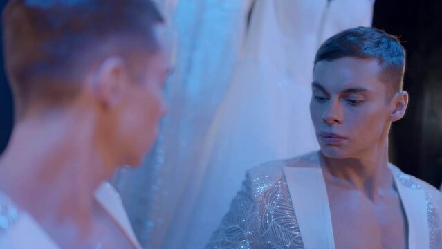 beautiful gay man in fashion jacket looking at mirror in atelier or cloth store, admiring reflection