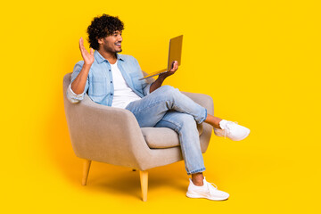 Full length photo of friendly man wear jeans shirt sit in armchair waving hand at laptop on video call isolated on yellow color background