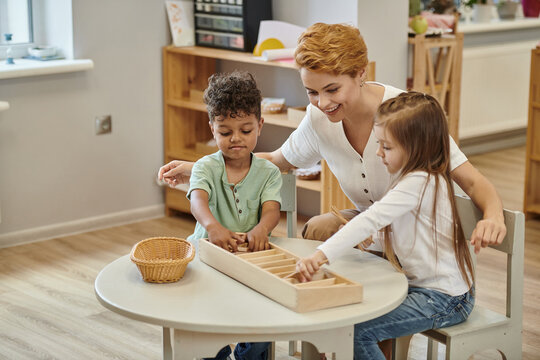 happy teacher talking to interracial kids playing with wooden didactic materials in montessori class