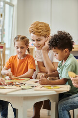 smiling teacher sitting near multiethnic kids playing with didactic material in montessori school - obrazy, fototapety, plakaty