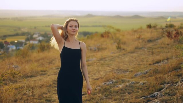 Beautiful slender young woman in a black dress is walking while turning to the camera circling on a meadow field outdoors at sunset, charmingly smiling and flirting