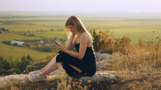 Young tourist girl sitting on the top of a rock and taking a photo of herself on a smartphone camera, beautiful landscape, outdoor walk in the countryside at sunset