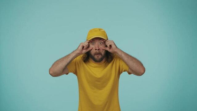 Delivery man in yellow uniform looking at the camera, making a big surprised eyes, head exploding gesture and amazed face expression. Isolated on blue background.
