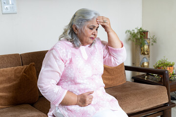 Tired, depressed senior woman sitting on couch in living room feeling hurt and lonely. Aged,...