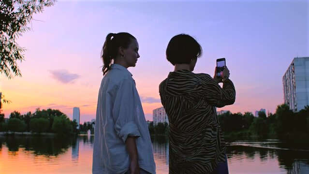Two women take photos of lake on camera. Beautiful girls taking video of sunset on mobile phone. Female couple friends in warm summer evening, nature landscape