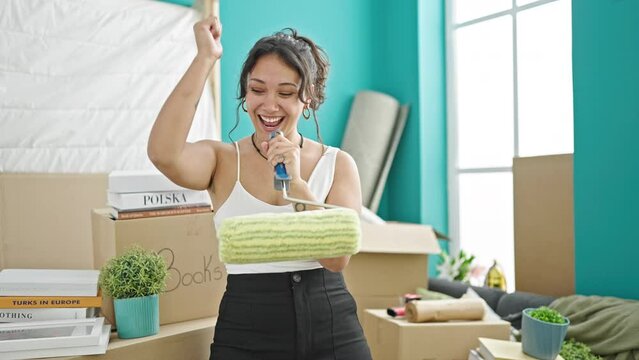 Young beautiful hispanic woman smiling confident singing song using paint roller as a microphone at new home