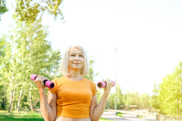 Senior woman going sport exercises with dumbbells on summer park. Active lifestyle concept.