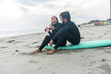 
A joyful surfer couple sits on a surfboard at the beach, sharing laughter and smiles as they enjoy the sun, sand, and each other's company in a perfect coastal moment.