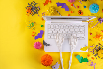 Funny high-colored Halloween bright yellow composition. White laptop with typing hands, Halloween decor colorful holiday accessories - spiders, cobwebs, pumpkins, bats, top view flatlay copy space