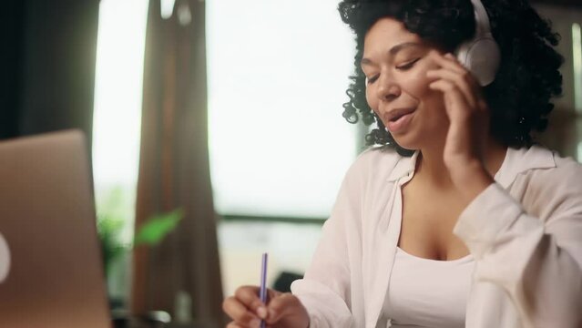 Pretty young woman freelancer with headphones listening music while working online typing browsing on laptop make notes and singing indoors Positive female enjoying great working day at cozy home