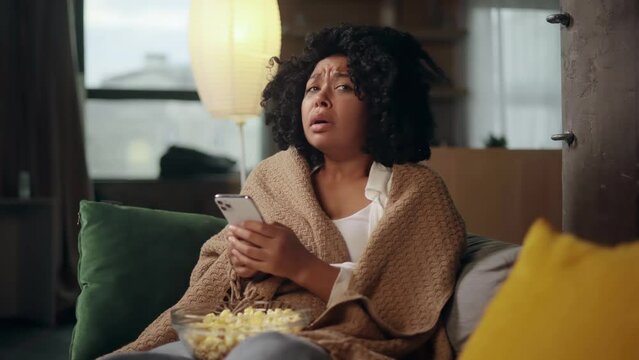 Portrait of crying young curly woman with smartphone sitting on the couch under the blanket and reading bad news email break up message from ex-boyfriend or personal problems at home	
