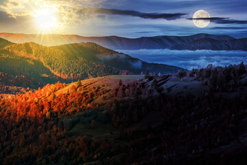 cold fog in the rural valley of Carpathian mountain range with sun and moon at twilight. trees with colorful foliage on the hillside meadow in morning light. day and night time change concept
