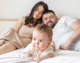 Fototapeta premium happy young family with baby infant kid boy sitting on bed, cuddling kissing child on hand, cheek, caresses. traditional family lying on bed at home.woman man kid portrait,posing in bedroom