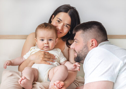 happy young family with baby infant kid boy sitting on bed, cuddling kissing child on hand, cheek, caresses. traditional family lying on bed at home.woman man kid portrait,posing in bedroom