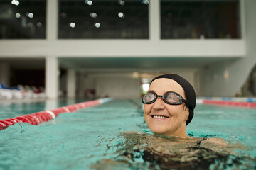Obraz premium cheerful middle aged woman in swim cap and goggles swimming in pool, water, recreation center, spa