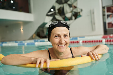happy middle aged woman in swim cap and goggles swimming with pool noodle, healthy lifestyle, sport
