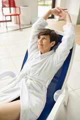 joyful middle aged woman with short hair stretching on lounger, white robe, spa center, smile