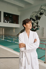 middle aged woman with short hair standing with folded arms, wearing white robe near pool, spa day