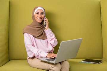 Young beautiful Muslim woman in hijab talking on phone use laptop to work in coworking space. Photo...
