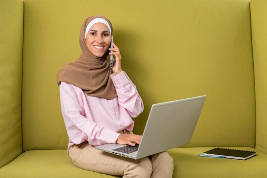 Young beautiful Muslim woman in hijab talking on phone use laptop to work in coworking space. Photo headshot. Smiling Arabian lady sitting on green sofa in free office place. Concept of study or work