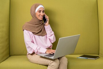 Young beautiful Muslim woman in hijab talking on phone use laptop to work in coworking space. Photo headshot. Smiling Arabian lady sitting on green sofa in free office place. Concept of study or work