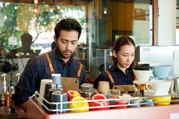 Beautiful Asian woman with Hispanic man concentrating on making orders that the customer has ordered intentionally. It's at the counter with a coffee machine at their business cafe.